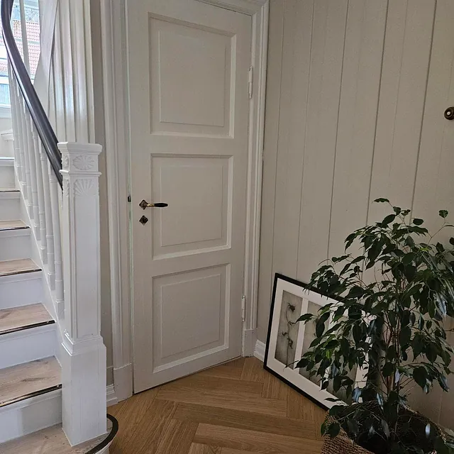 Lys gang med hvit innerdør, trapp med detaljer og plante i flettet kurv på tregulv.