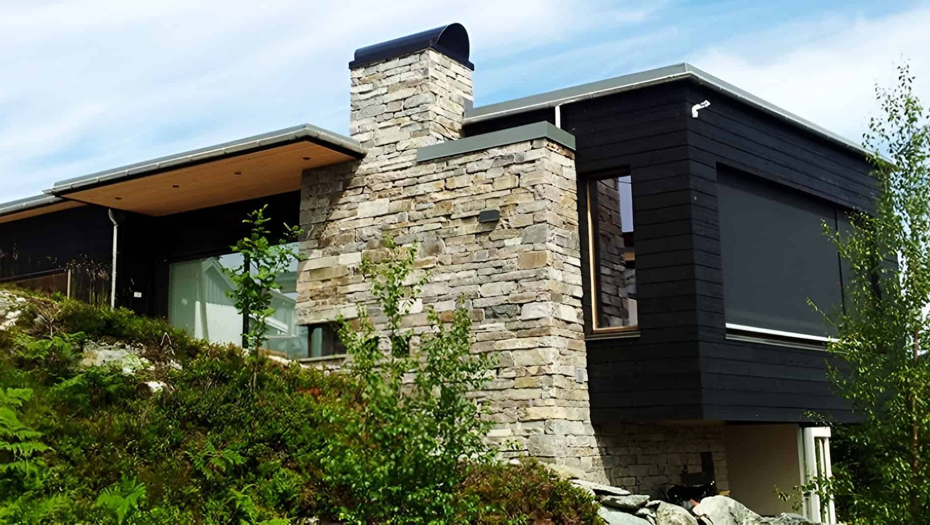 Hus med stein og mørk kledning, store vinduer og flatt tak på en grønn åsside