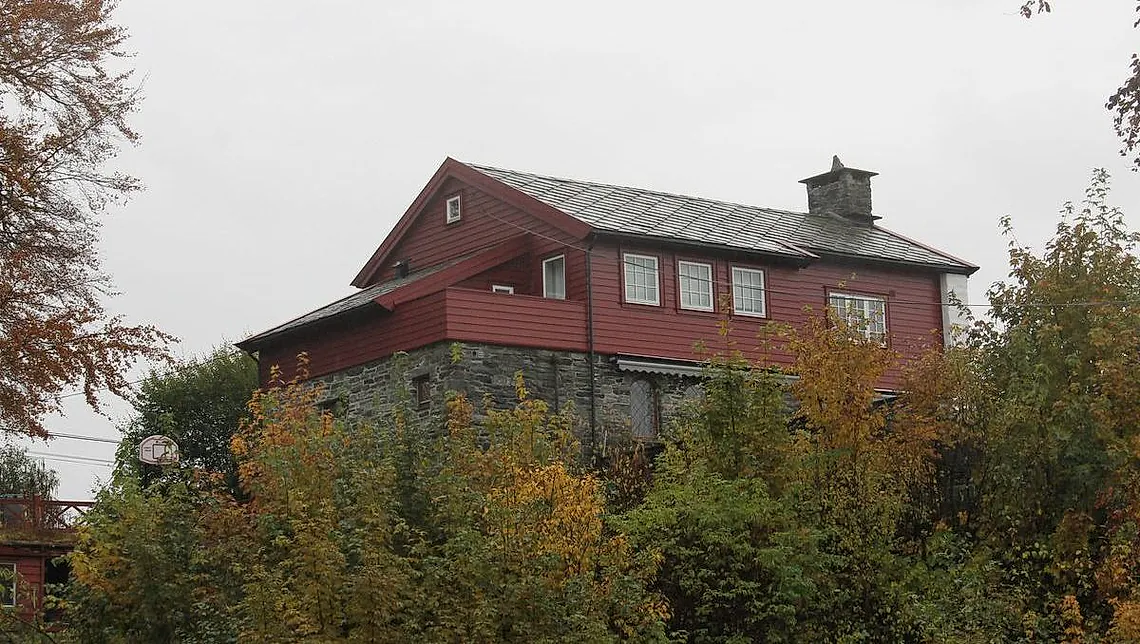 To-etasjes hus med rød fasade og grått tak, omgitt av høstfarget vegetasjon