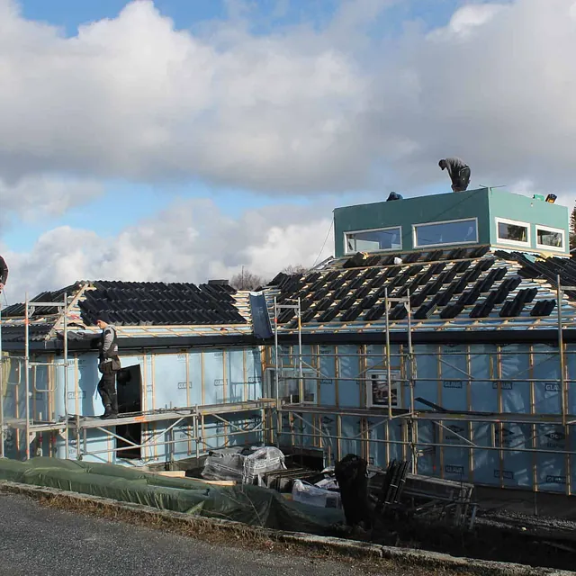 Hus under bygging med blå isolasjonsplater og arbeidere som legger takstein, omgitt av stillas.